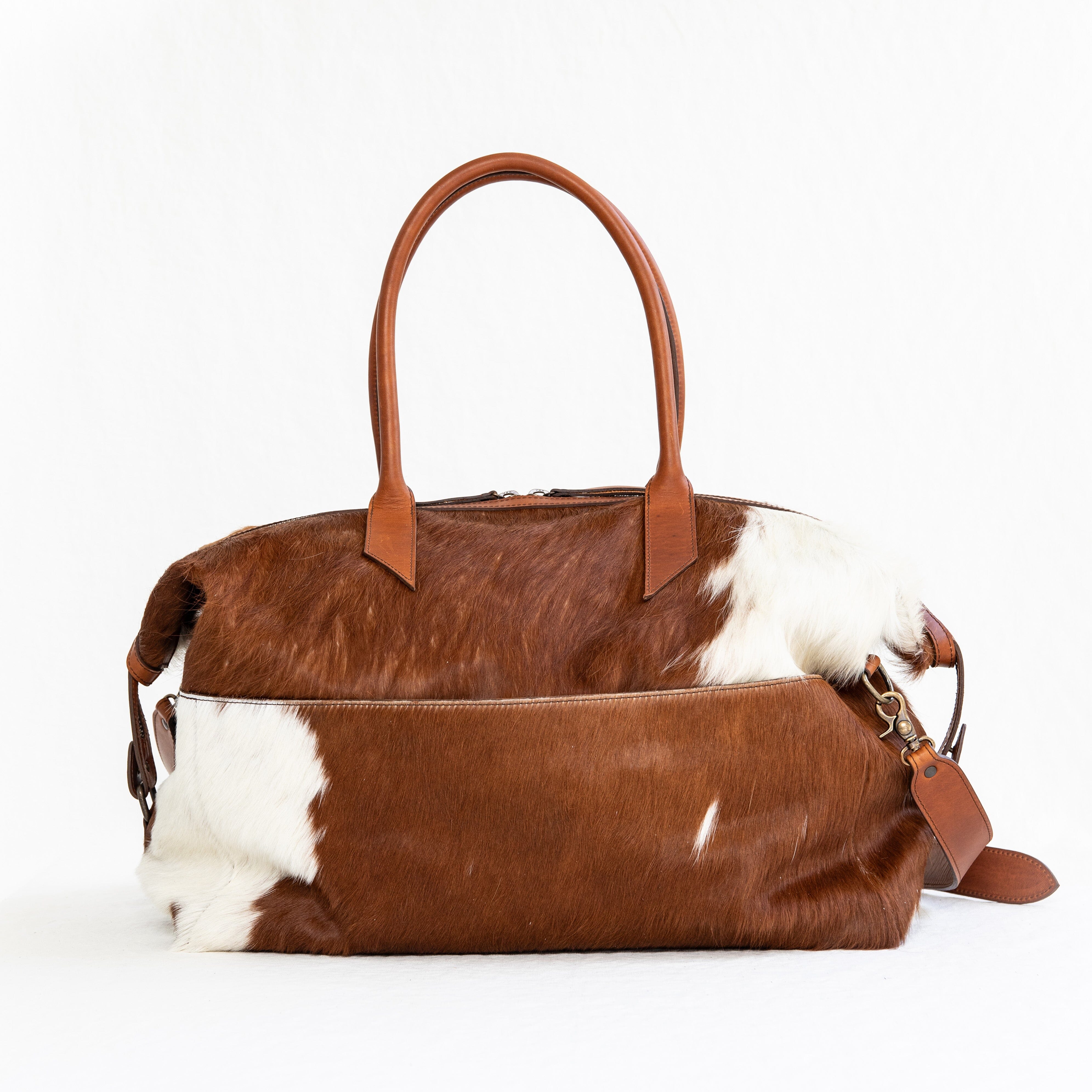A brown duffle bag with a hair-on-hide design, featuring a leather handle and shoulder strap, brass clasps, and zipper enclosures.