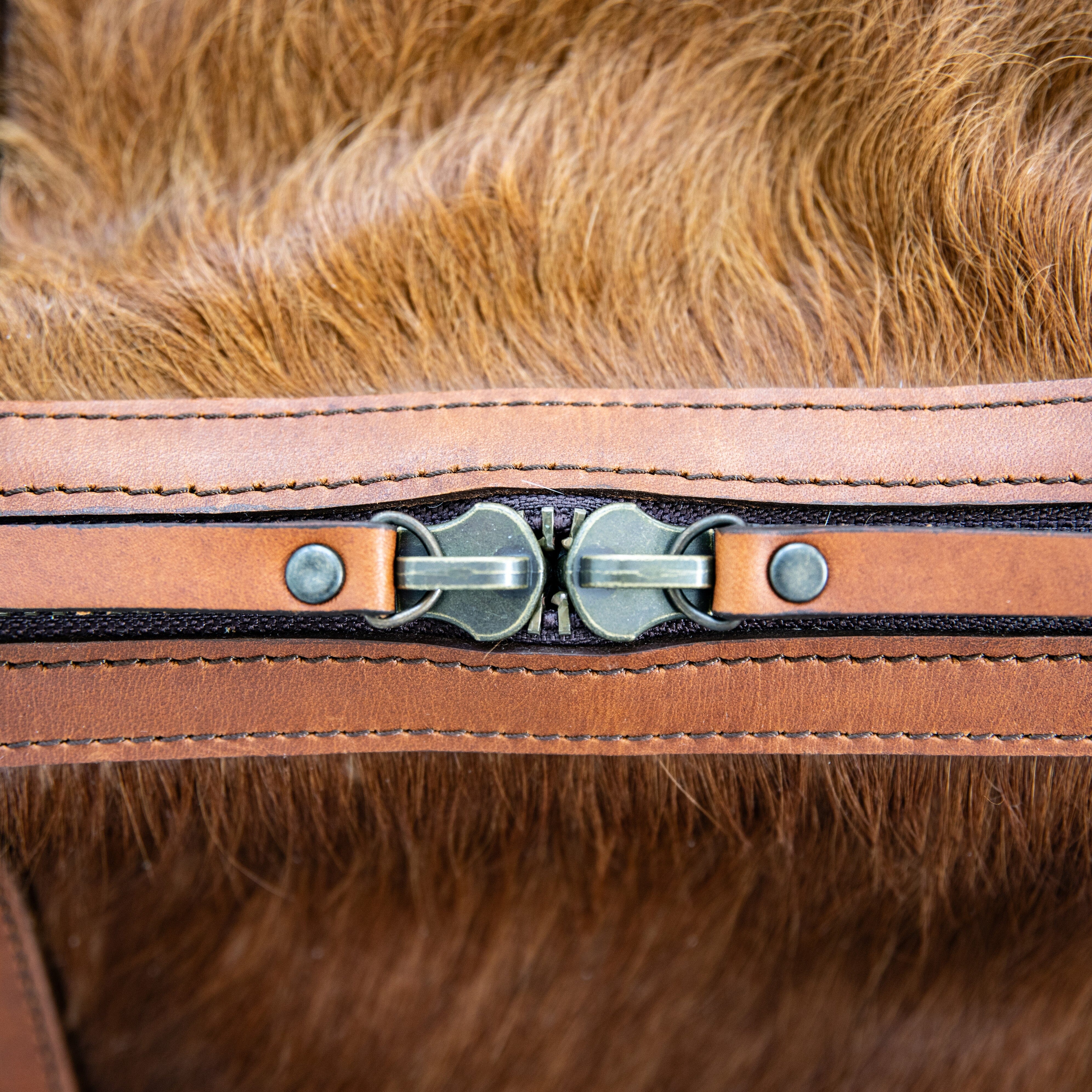 A brown duffle bag with a hair-on-hide design, featuring a leather handle and shoulder strap, brass clasps, and zipper enclosures.