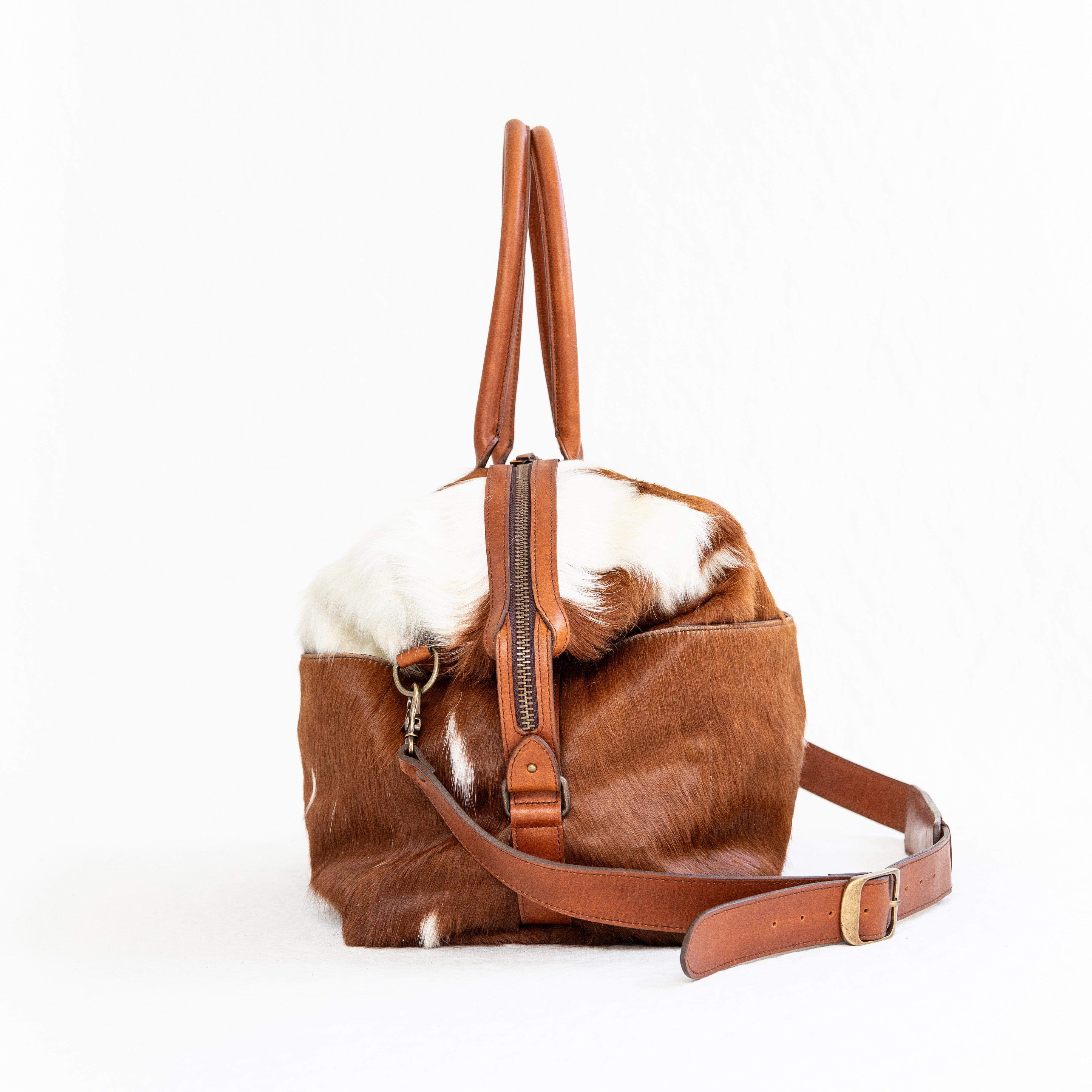 A brown duffle bag with a hair-on-hide design, featuring a leather handle and shoulder strap, brass clasps, and zipper enclosures.