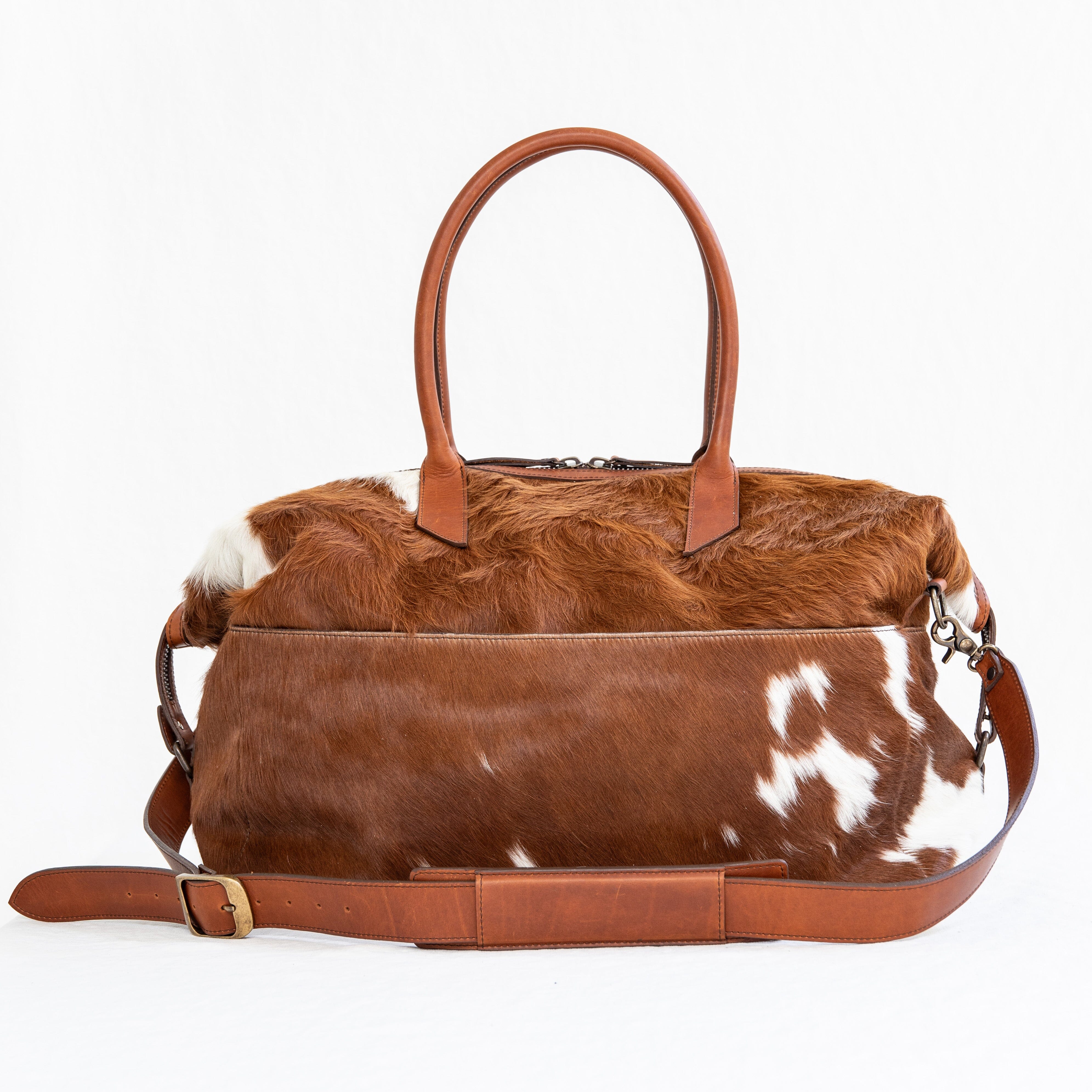 A brown duffle bag with a hair-on-hide design, featuring a leather handle and shoulder strap, brass clasps, and zipper enclosures.