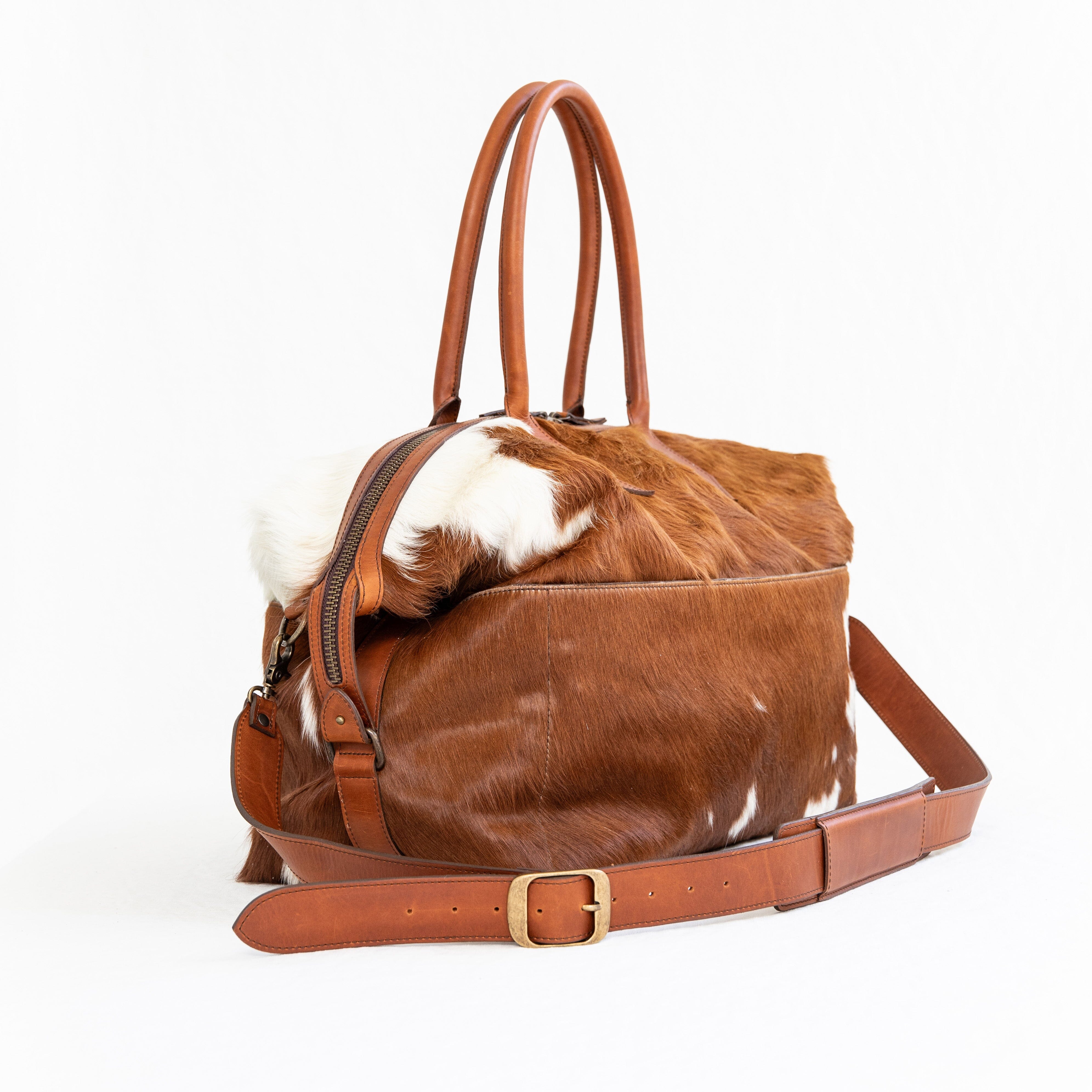 A brown duffle bag with a hair-on-hide design, featuring a leather handle and shoulder strap, brass clasps, and zipper enclosures.