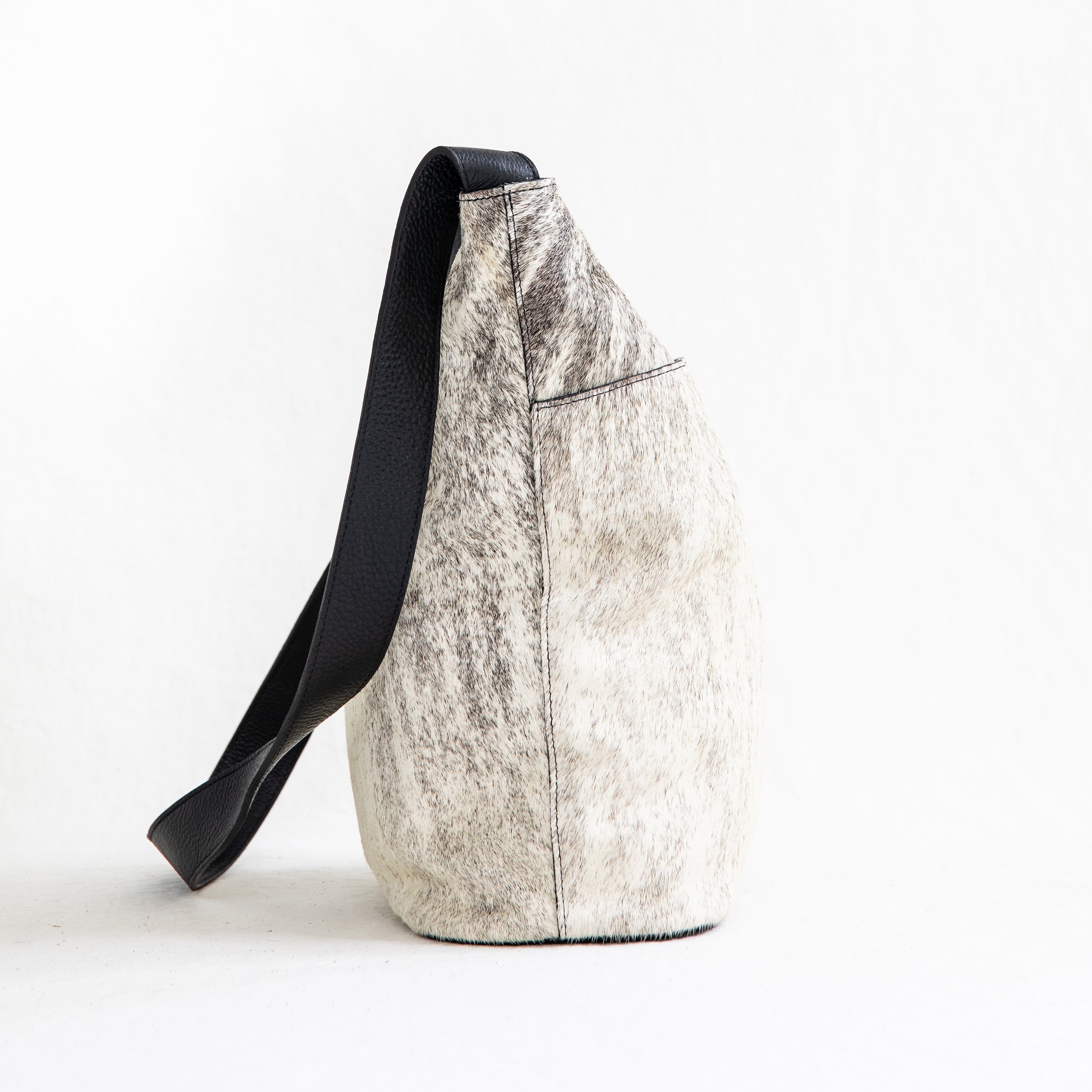 A white and beige crossbody shoulder bag made of hair-on-hide leather, featuring a brass clasp and a black shoulder strap.